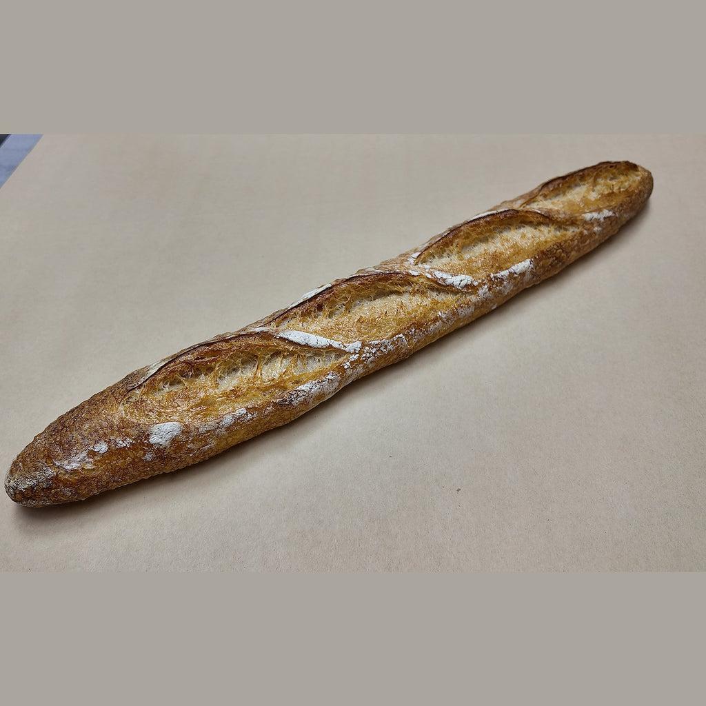 A single rustic baguette with a golden-brown crust and dusting of white flour, placed diagonally on a plain beige surface.