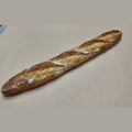 A single rustic baguette with a golden-brown crust and dusting of white flour, placed diagonally on a plain beige surface.