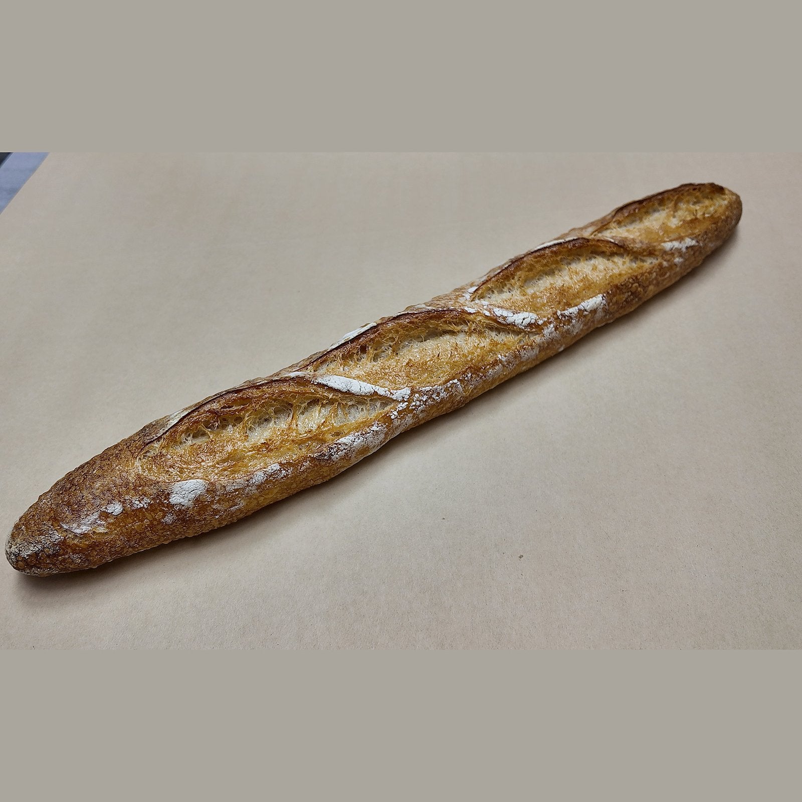 A single rustic baguette with a golden-brown crust and dusting of white flour, placed diagonally on a plain beige surface.